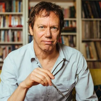 Portrait of author Robert Greene seated in front of a bookshelf, holding his glasses